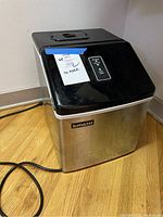 View of the entire ice maker showing its black plastic top and stainless steel body with power cord extended on wooden floor.