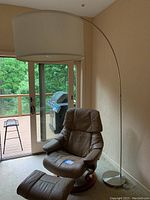 Full view of lamp beside leather chair showing long arched nickel finish metal frame and large white fabric lampshade.