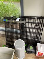 Two large folded black metal wire dog exercise pens leaning against a wall by a window, with a white and grey Little Champ cat litter disposal container and other pet waste accessories on the floor.