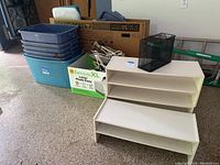 Photo showing six stacked Rubbermaid storage bins, a box of hangers, and three white shoe shelves, along with the black wire mesh box on top of one shelf.