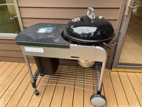 Full view of Weber charcoal kettle grill mounted on rolling cart with side table, charcoal bin, bottom wire shelf, and wheels