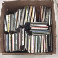 Photo of a cardboard box filled with numerous CD jewel cases arranged upright showing the spines and some covers visible