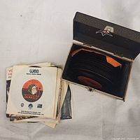 Top view showing a stack of 45 RPM vinyl records inside a black textured storage box, with more records stacked outside on the surface.