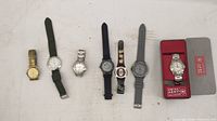 Seven assorted wristwatches displayed side by side including various colors and strap materials; some metal bands, one in box