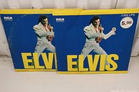 Two Elvis Presley vinyl record albums standing upright showing cover art of Elvis in white jumpsuit on blue background with yellow text.