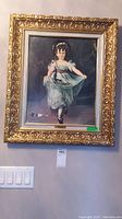 Framed art of a young girl dressed in a Victorian dress and bonnet, holding dress edges, ornate gold frame visible.