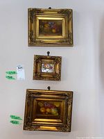 Three framed framed paintings on wall: Two larger paintings with fruit still life and one smaller floral painting in ornate gold frames.