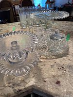 Two ribbed glass cake plates with ruffled edges in front, glass trays and etched sugar, creamer, dispenser set at back on countertop.
