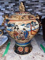 Front view of ornate Asian jar showing detailed hand-painted scene of traditionally dressed figures near a boat, gold lion head handles, and decorative pedestal base.