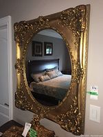 Front view of the ornate gold resin framed wall mirror hung on a wall reflecting a bed and window.