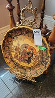 Photo showing round decorative Asian ceramic plate on ornate gold-toned metal stand, placed on floor near wooden chair and black tiled floor.