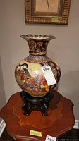 Full view of the decorated vase on its wooden stand, showing intricate painted scene with multiple people and garden elements