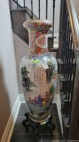 Tall Chinese floor vase with scenic hand-painted figures and calligraphy shown upright on wooden stand near staircase.