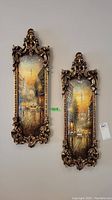 Two vertically oriented framed paintings showing Paris street scenes with Eiffel Tower, in decorative gold ornate frames.