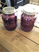 Photo shows two matching purple glass jars with wire hanging handles and silver metal lids on a wooden surface.