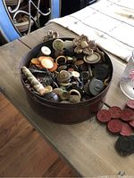 Tin filled with various antique buttons of different sizes, colors, and materials, including some with floral designs, along with red and black checkers pieces on the table beside the tin.