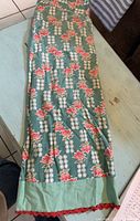 Long vintage apron with green base and red floral pattern, with red ruffled hem, fully laid out on table.