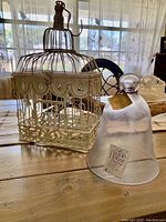 Photo showing the decorative metal bird cage with scalloped trim alongside a clear glass bell jar cloche with label, placed on a wooden table.