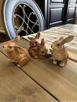 Three ceramic bunnies on wooden table, detailed textures and natural brown coloring.