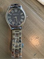Close-up of men's watch showing silver-tone case, black dial with Roman numerals, crystal accents on the case edges, and metal link bracelet.