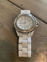 Creamy white women's ceramic watch resting on wooden surface, face visible showing clear numerals and silver scalloped bezel.