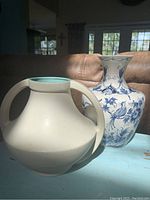 Two vases on a blue table: white Coors Colorado pottery vase with rounded handles, and blue and white hand-painted Blue Delfts vase with floral and bird motifs.