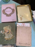 Pink 'Rendezvous' stationery box open with assorted paper sheets inside and a green 'Forget-me-not Stationery' box with illustration of little girl holding flowers.