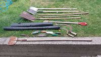 View of assorted garden tools laid on grass including shovels, sickles, hoe, and garden fabric rolls.
