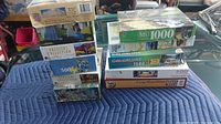 Photo showing stack of assorted puzzle boxes with various brands and piece counts from 500 to 1500 pieces, placed on padded blue blanket