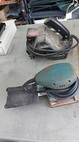 Photo showing two sanders on a table, dusty Craftsman belt sander and teal Mechanics palm sander coiled cords