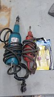Two electric die grinders (one blue Makita, one red) with coiled cords and a set of grinding accessories in blister pack.