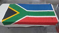 A full view of the South African flag spread out on a white surface showing the various colors and some dirt marks on the fabric.