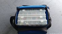 Blue soft fishing tackle bag with three stacked clear plastic tackle boxes on top showing compartments with lures.