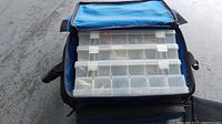Blue soft tackle bag with multiple transparent plastic compartment boxes inside, organized for fishing gear storage