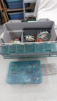Large gray tackle box open showing fishing lines, lures, and bobbers inside with transparent green lid compartments