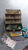 Closed view of the tackle box with contents and fishing accessories laid in front showing various lures, weights, and packages