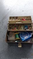 Open metal tackle box with three-tiered compartments filled with assorted fishing lures, hooks, bobbers, and fishing line.