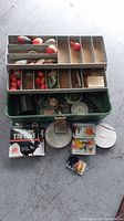 Green metal tackle box open showing fishing gear inside with bobbers and lures visible