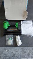 Photo showing the Kawasaki 18 gauge brad nailer and stapler in green and black, oil bottle, gloves, instruction manual, and original box.