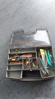 Plastic tackle box open showing various metal and plastic lures with hooks and fishing line inside