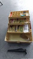 Beige tackle box opened showing three tiered trays with various fishing lures, hooks, and rigs inside compartments.
