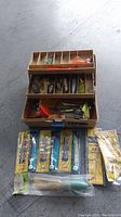 Overview photo showing opened tackle box filled with assorted saltwater fishing gear including lures and sinkers, with multiple packaged fishing lures laid out in front.