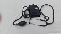 Photo showing vintage black sphygmomanometer cuff, inflation bulb, and black stethoscope on white surface.