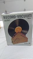 Photo of the front of the original box showing the Ronco Record-Vacuum, cordless electric device cleaning a vinyl record.