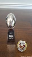 Miniature Vince Lombardi Trophy next to Patrick Mahomes II replica Super Bowl ring shown on wood floor.