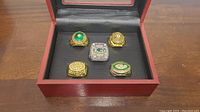 Five Green Bay Packers replica Super Bowl rings displayed in a wooden box with black velvet interior, showing detailed designs and stones.