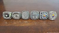 All six Pittsburgh Steelers replica Super Bowl rings displayed side by side on a wooden surface, showing various styles and designs.