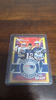 Gold rookie card featuring Tom Brady in New England Patriots uniform in a plastic protective case on a wooden surface.