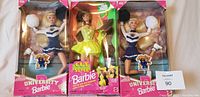 Front view of three Barbie dolls in original packaging: two University Barbie Kentucky Cheerleader dolls in blue and white uniforms, one Cut & Style Barbie doll in yellow outfit with styling accessories.