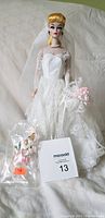 Full view of Barbie Wedding Party Porcelain Doll in white lace wedding dress with bouquet and additional flowers accessory bag, showing doll standing on bed.